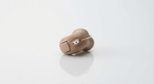 Microscopic custom-crafted in-the-ear invisible Oticon Own hearing aid shell designed to fit discreetly inside human ear canal photographed on minimalist white reflective table.